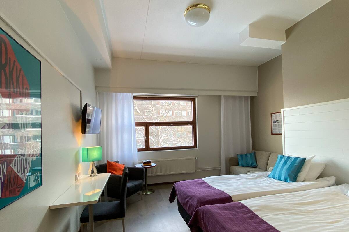Hotellihuone, jossa on kaksi sänkyä, työpöytä, tuoli ja ikkuna, josta on näkymä rakennuksiin.