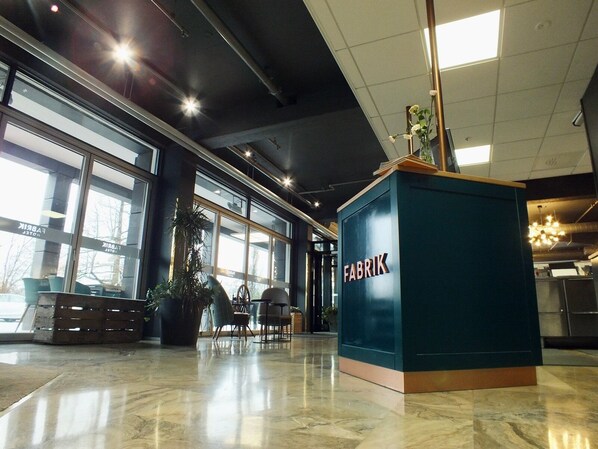 A modern lobby with a large blue counter labeled "FABRIK," large windows, and a polished marble floor. - Hotel Fabrik Forssa (Forssa)