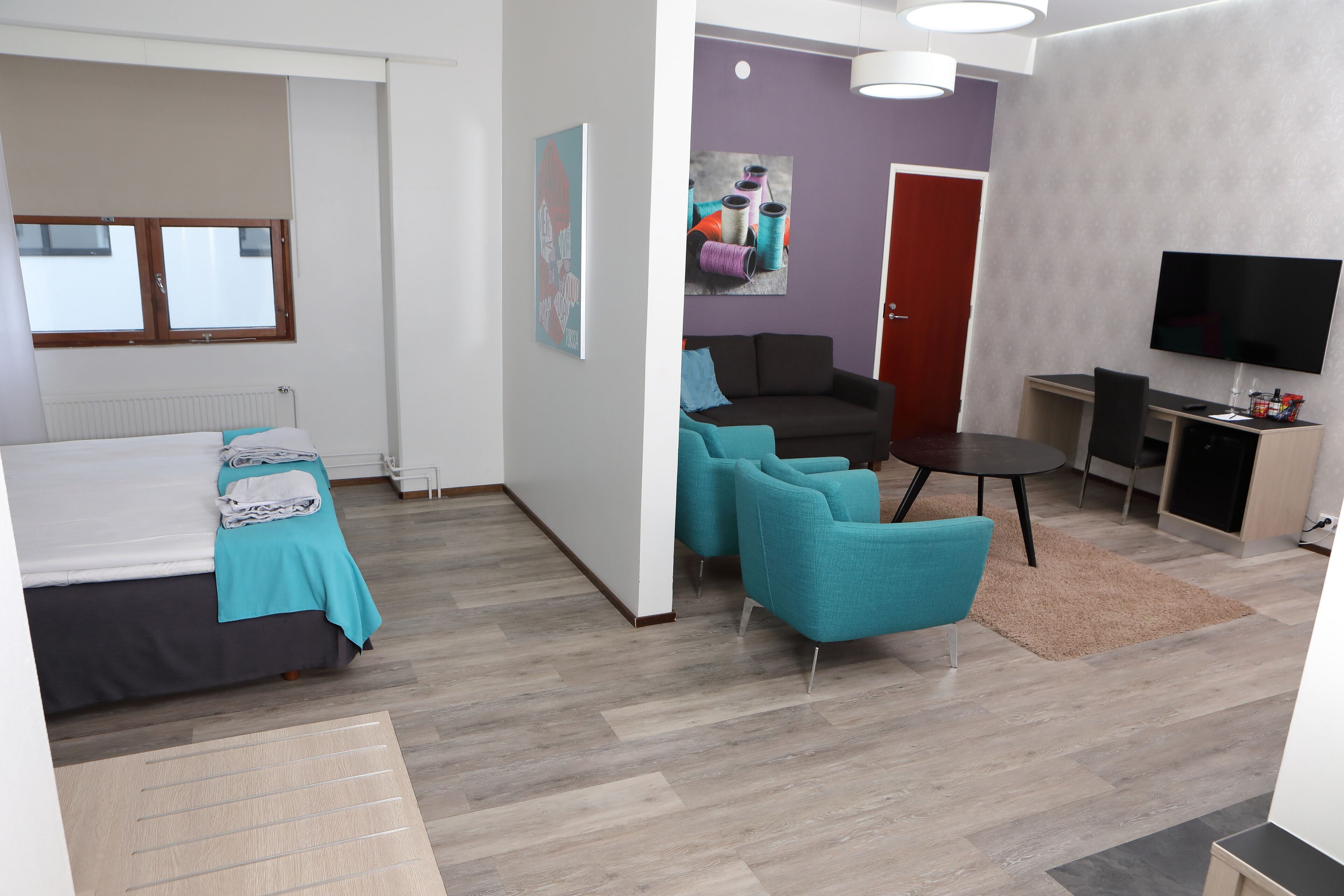 A modern hotel room with a bed, a desk, a TV, and teal chairs.