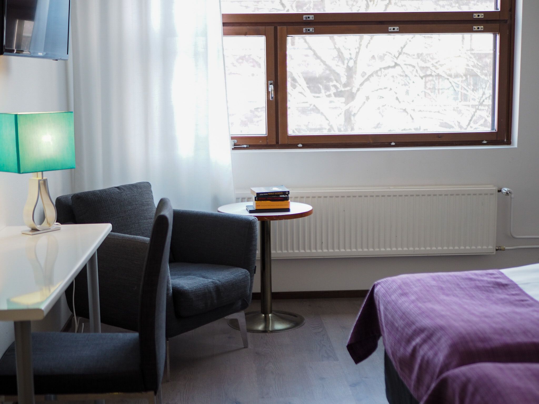 Habitación de hotel con cama, escritorio con lámpara, silla, pequeña mesa con libros y ventana con vista a árboles.