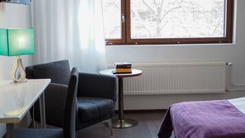 Habitación de hotel con cama, escritorio con lámpara, silla, pequeña mesa con libros y ventana con vista a árboles.