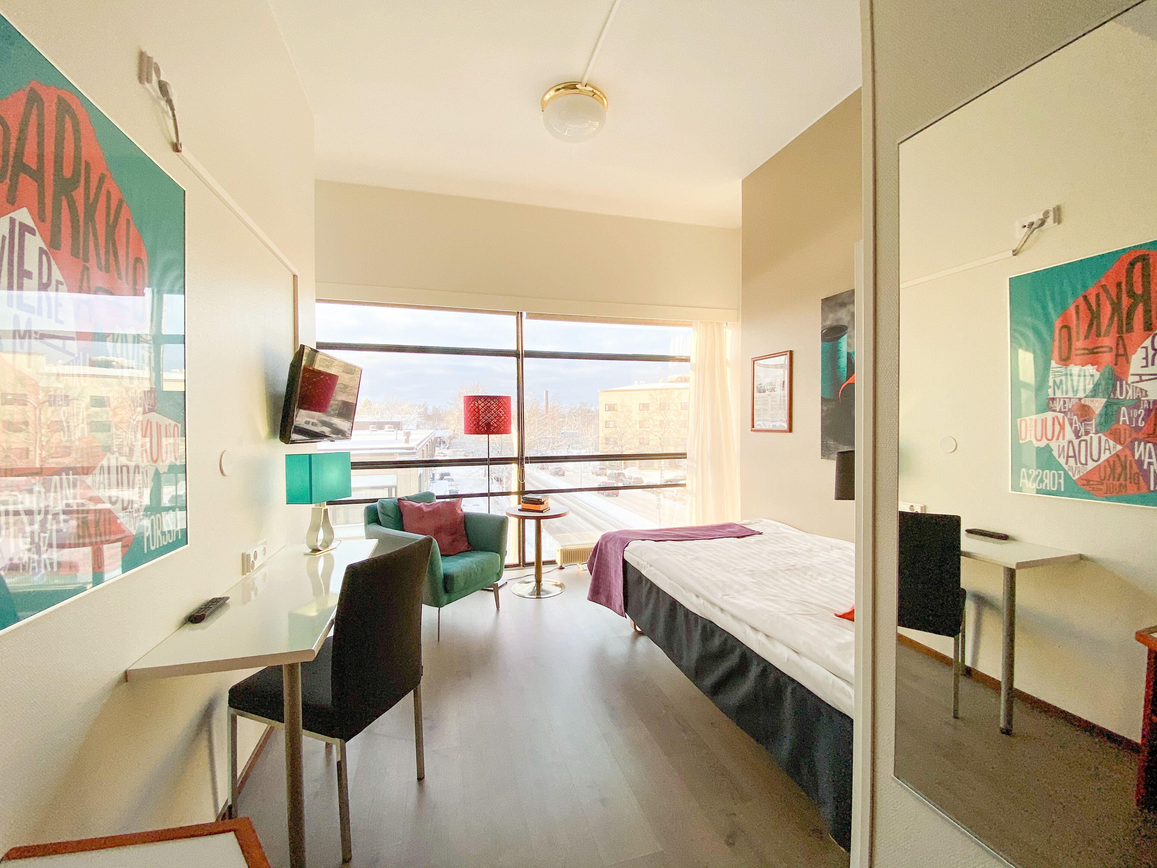 A modern hotel room with a large window, a desk, a chair, a bed, and a small table.