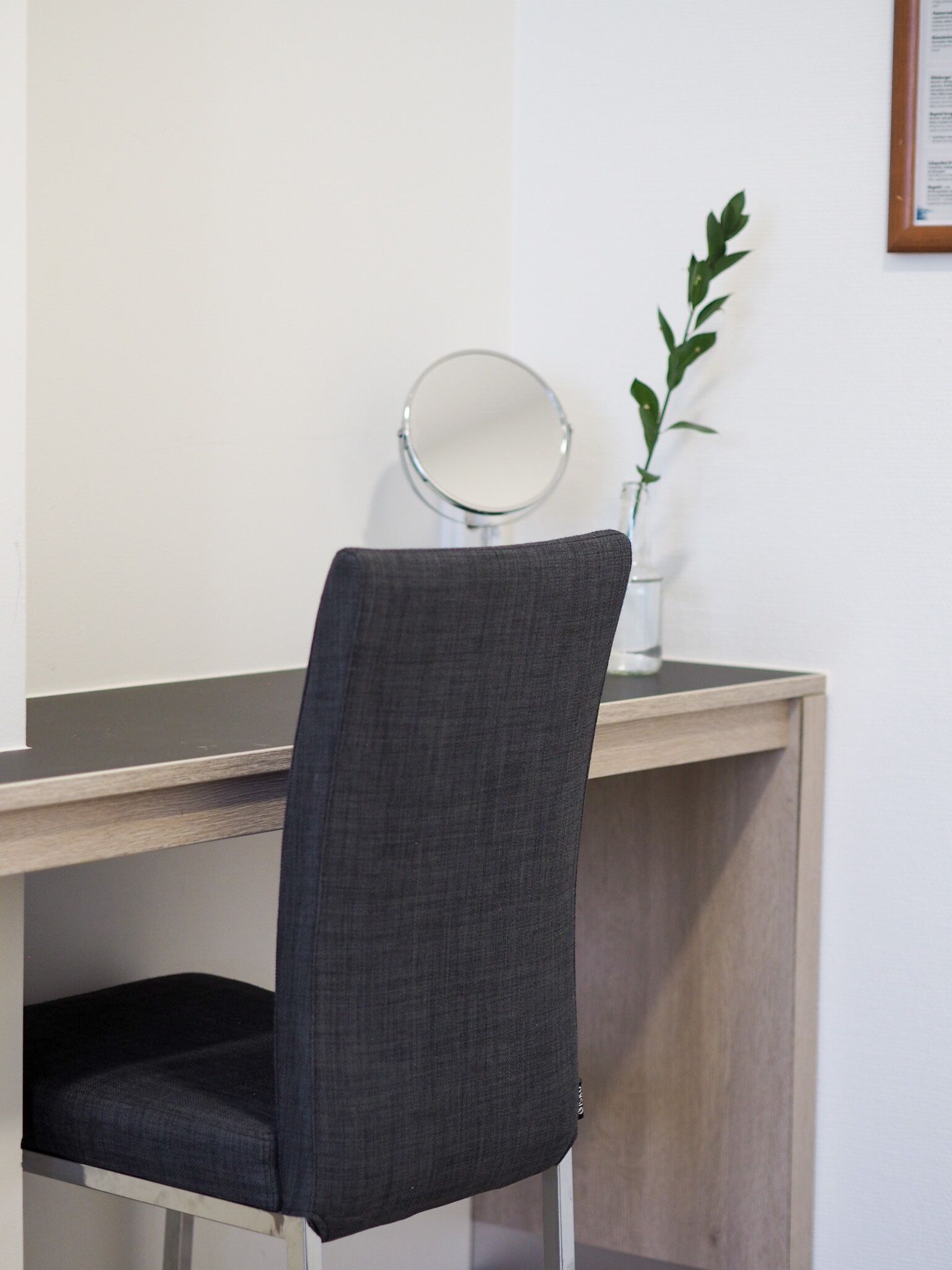 Una silla con respaldo de tela oscura y asiento acolchado, ubicada frente a un escritorio con un espejo redondo y una pequeña planta en maceta.