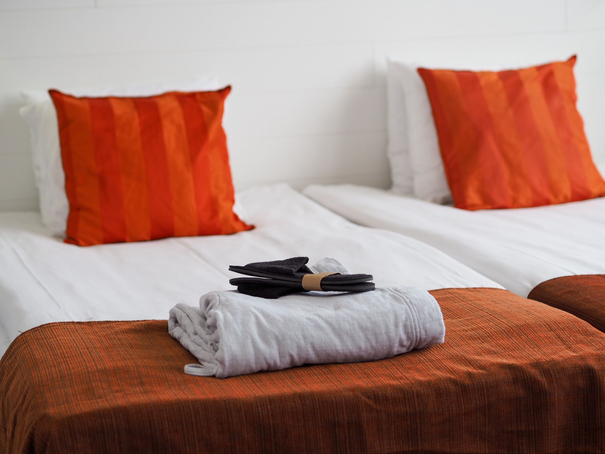 Quarto de hotel com duas camas, cada uma com uma almofada laranja e roupa de cama branca. Há uma toalha dobrada e um objeto preto na cama em primeiro plano.