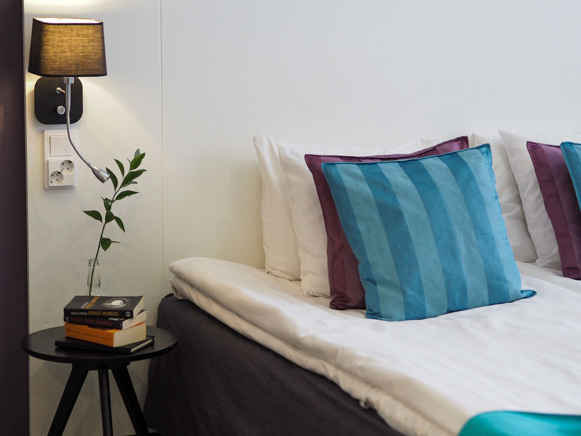 A neatly made bed with white and striped pillows, a bedside lamp, a small table with a plant and books, and a power outlet on the wall.