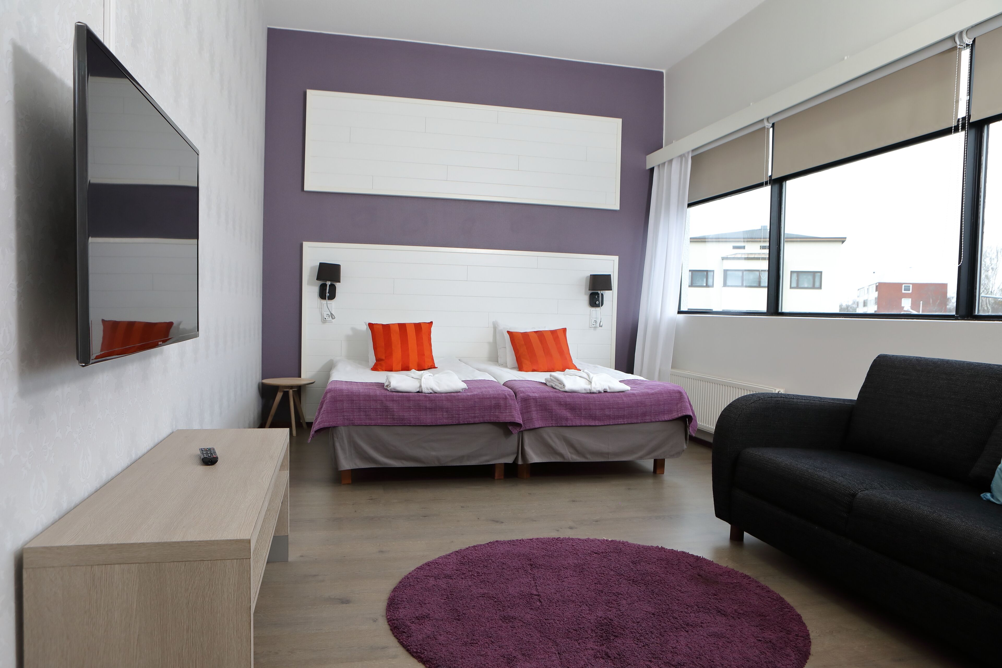 Una habitación de hotel moderna con una cama grande, un sofá, una mesita pequeña y un ventanal amplio.