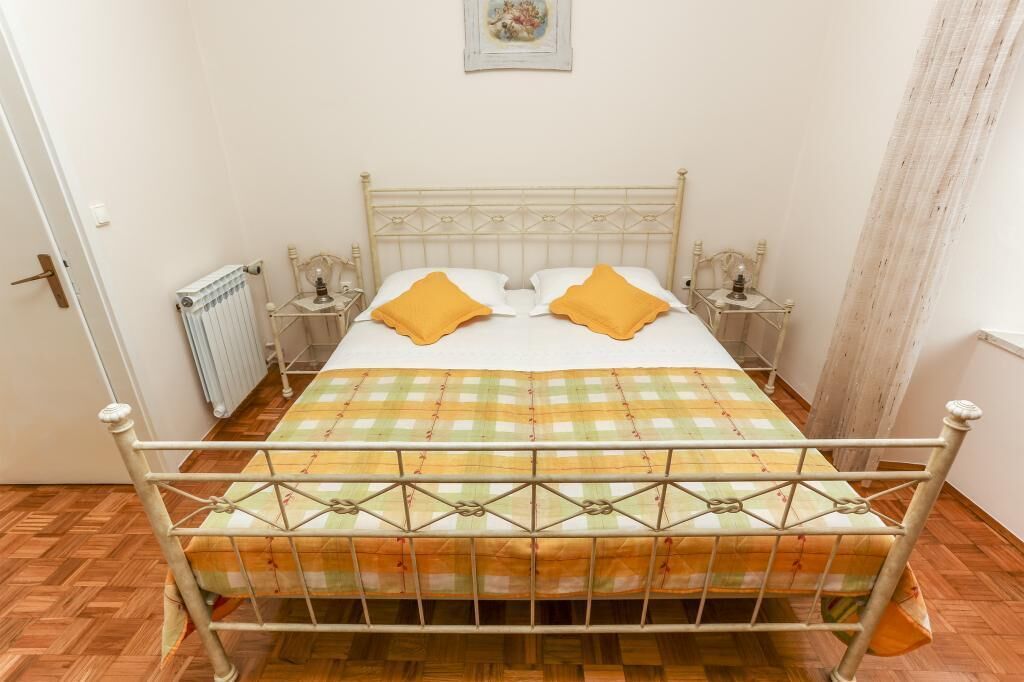 A bed with a metal frame, a checkered bedspread, and two pillows. There is a radiator on the wall and a small table with a lamp beside the bed.