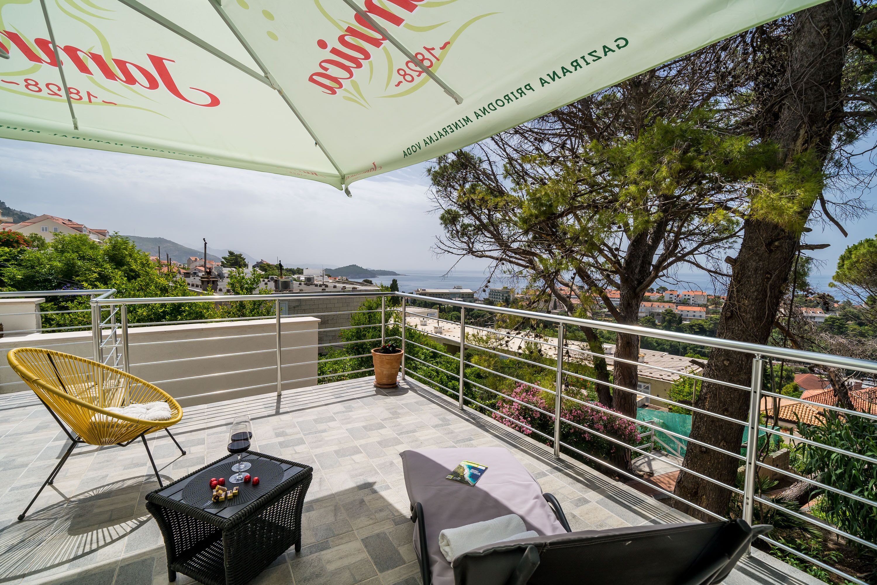 Eine Terrasse mit Gartenmöbeln, einem Tisch mit Flasche und Gläsern sowie Blick auf das Meer und Gebäude.
