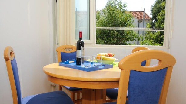 A small dining area with a round wooden table, blue cushioned chairs, a wine bottle, a glass, and a bowl of fruit. - Apartments Depozit-joky (Dubrovnik)