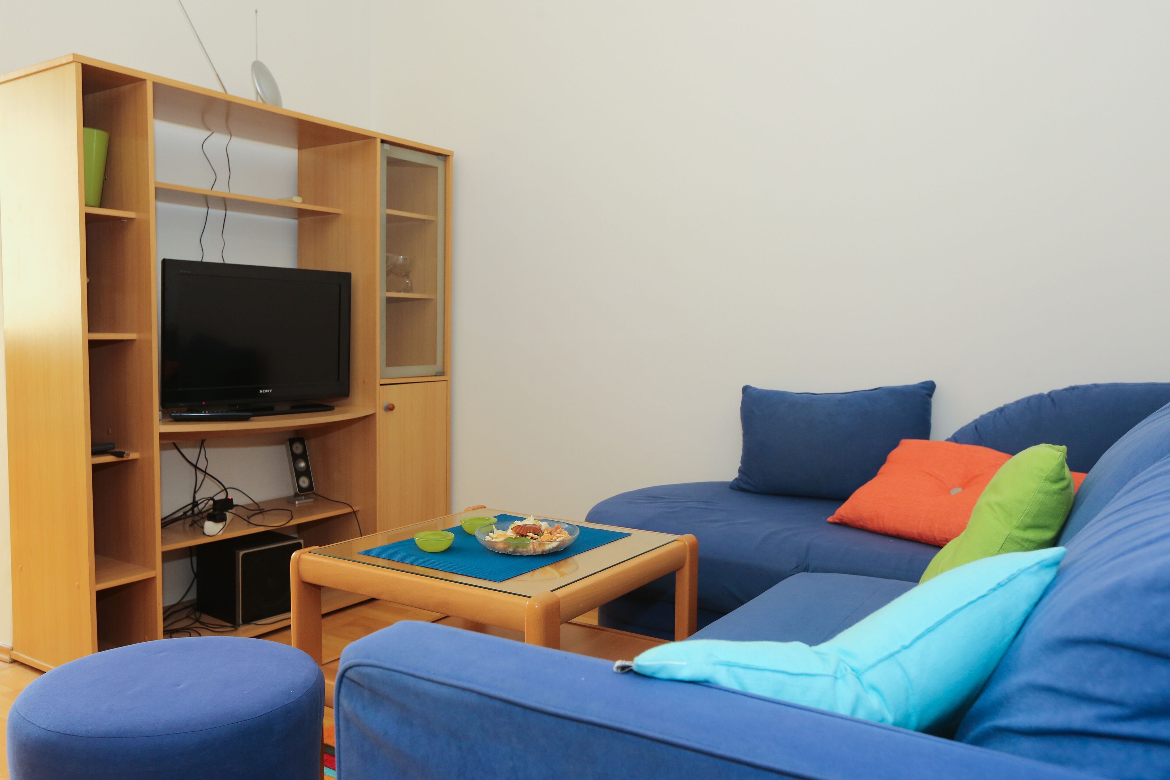 A living room with a blue sofa, a wooden entertainment unit with a flat-screen TV, and a small wooden table with a placemat.