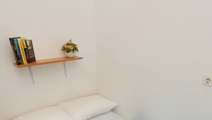 A neatly made bed with a blue bedspread and white pillows, a small shelf with books and a flower, and a wooden floor. - Apartments Depozit-joky (Dubrovnik)