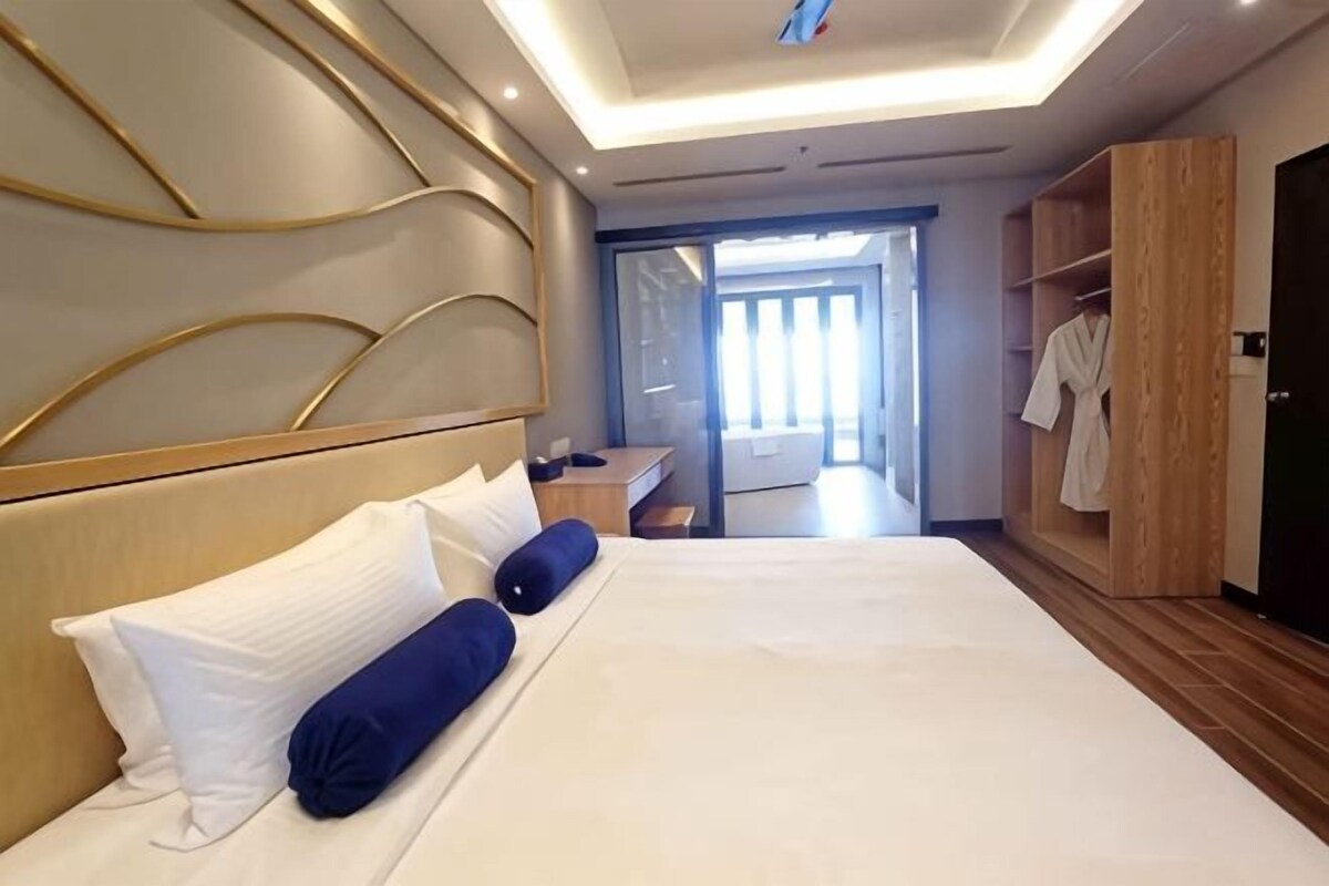 A hotel room with a large bed, a wooden headboard, a wardrobe, and a bathroom visible through a glass door.