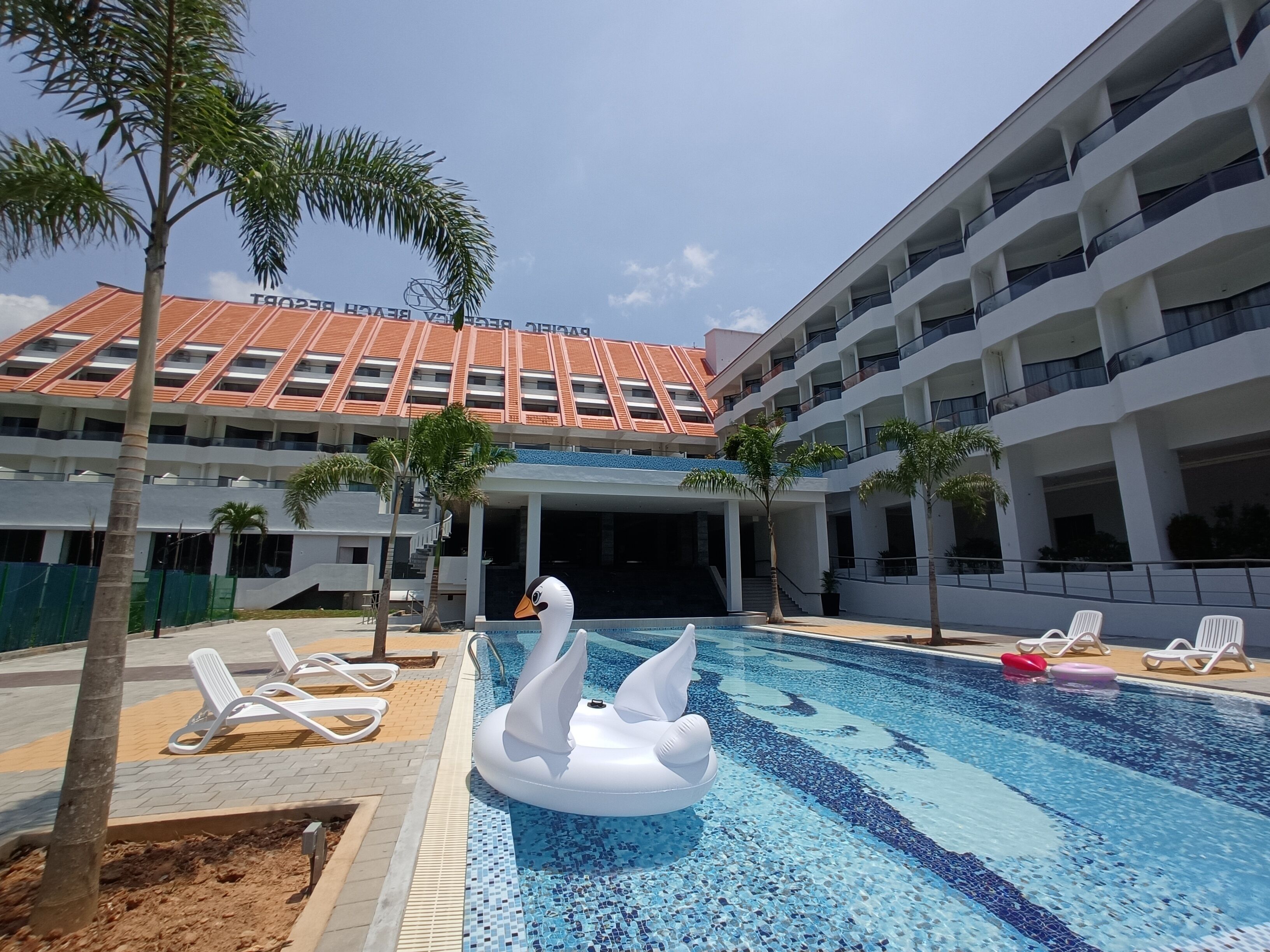 A swimming pool with a swan float, surrounded by lounge chairs and palm trees, with a building featuring a red-tiled roof in the background.