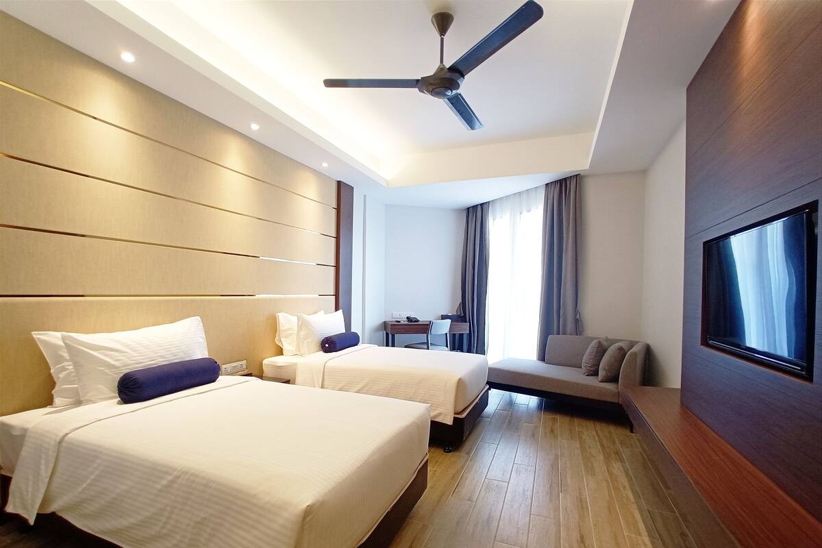 A hotel room with two beds, a sofa, a desk, a TV, and a ceiling fan.