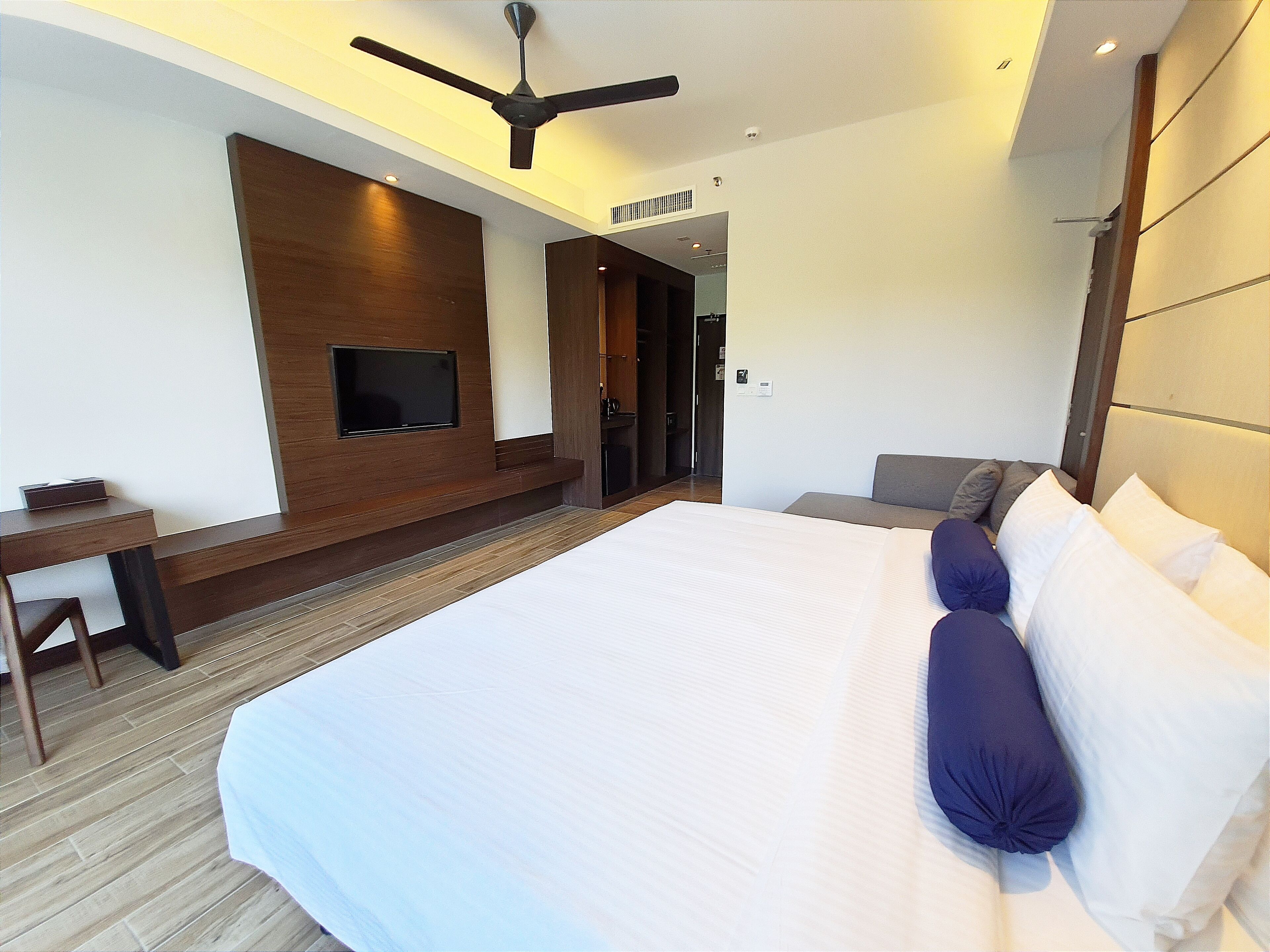 A modern hotel room with a large bed, a flat-screen TV mounted on the wall, a desk, and a ceiling fan.