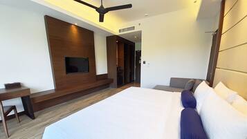 A modern hotel room with a large bed, a flat-screen TV mounted on the wall, a desk, and a ceiling fan.