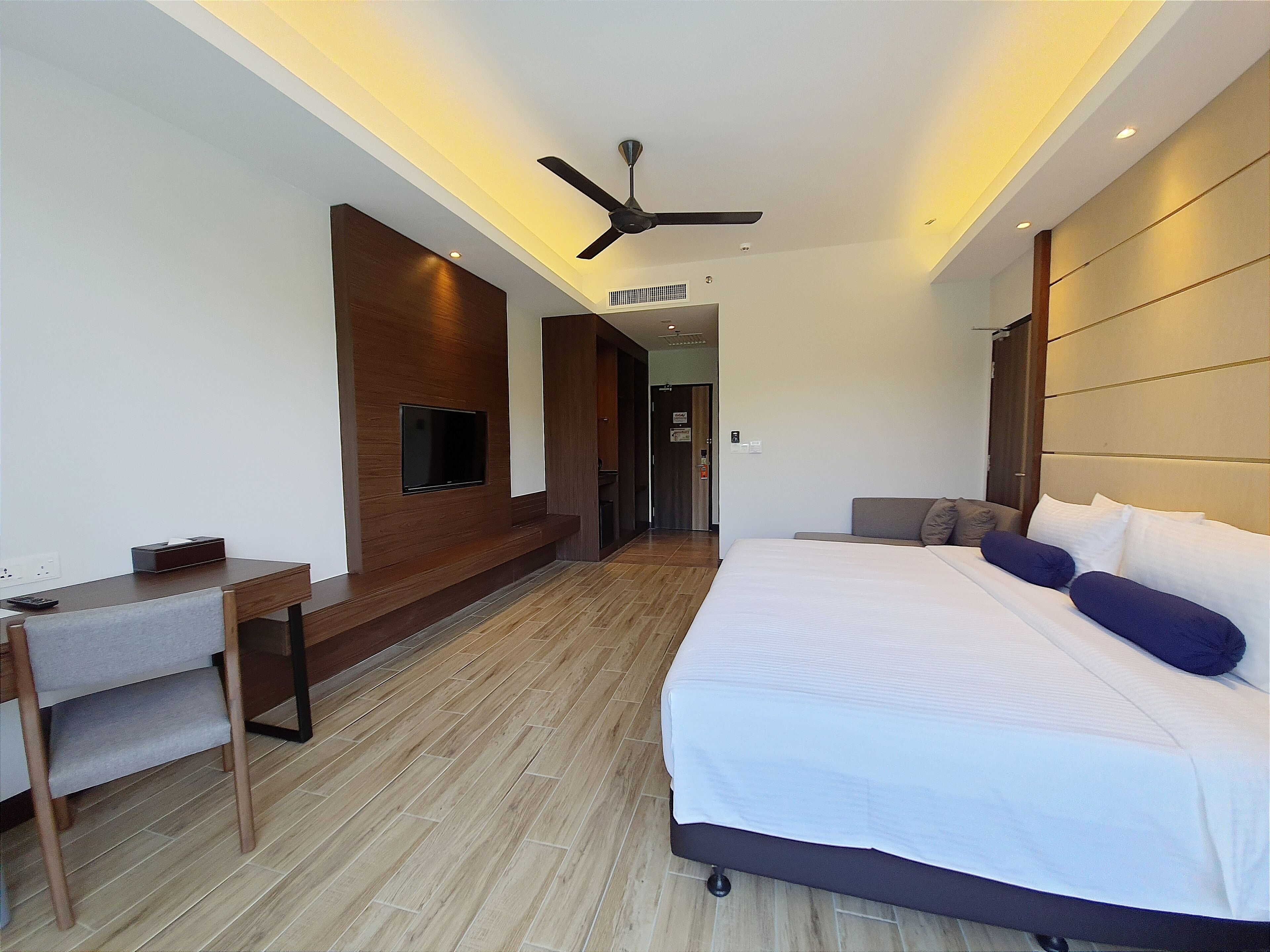 A modern hotel room with a large bed, a desk, a TV mounted on the wall, and a ceiling fan.
