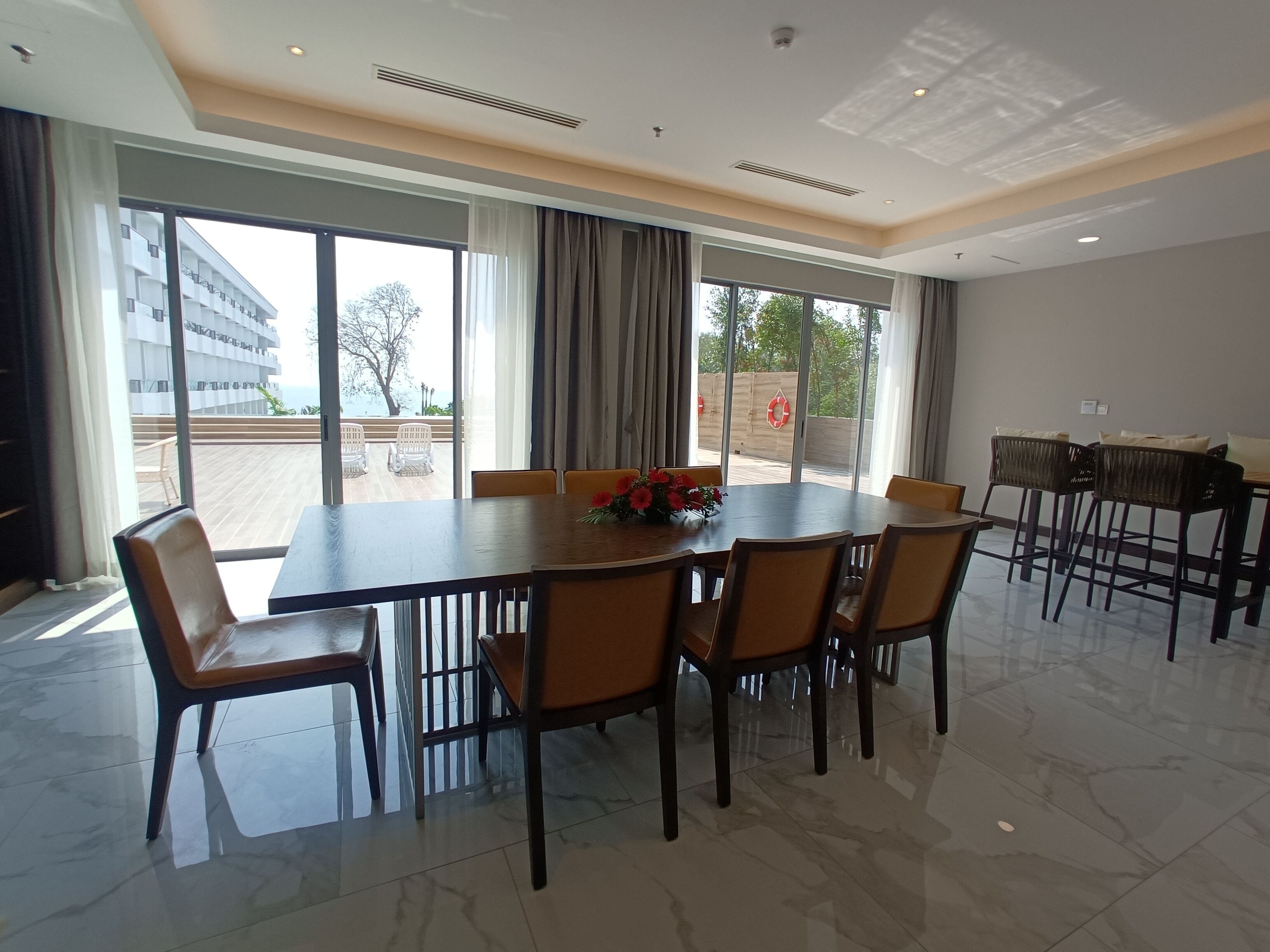 Un comedor amplio con una mesa grande y sillas, ventanas grandes con cortinas, y vista a un edificio y árboles al exterior.