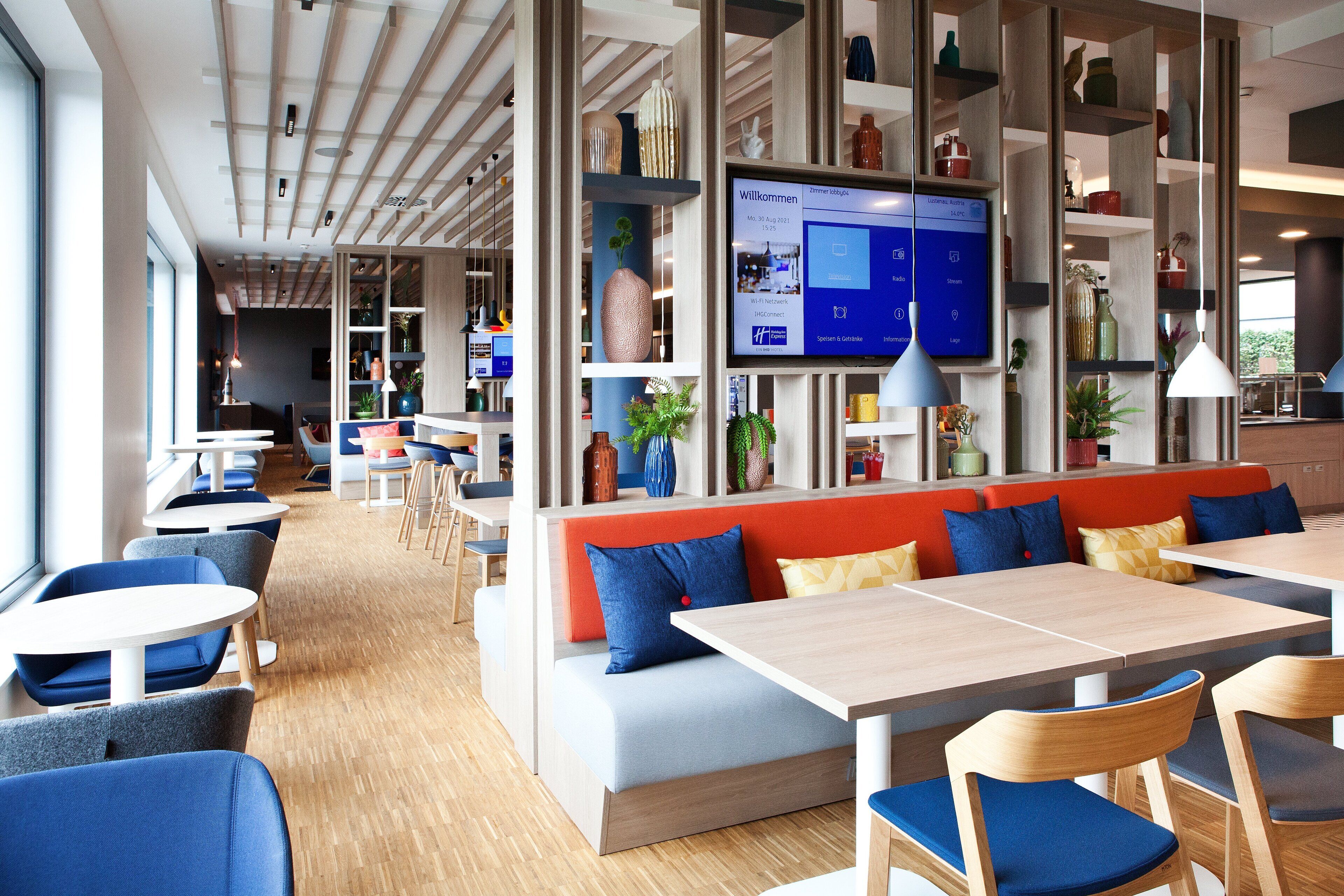 A modern restaurant with high tables and blue chairs, a long bench with orange cushions, and a TV screen displaying a menu.