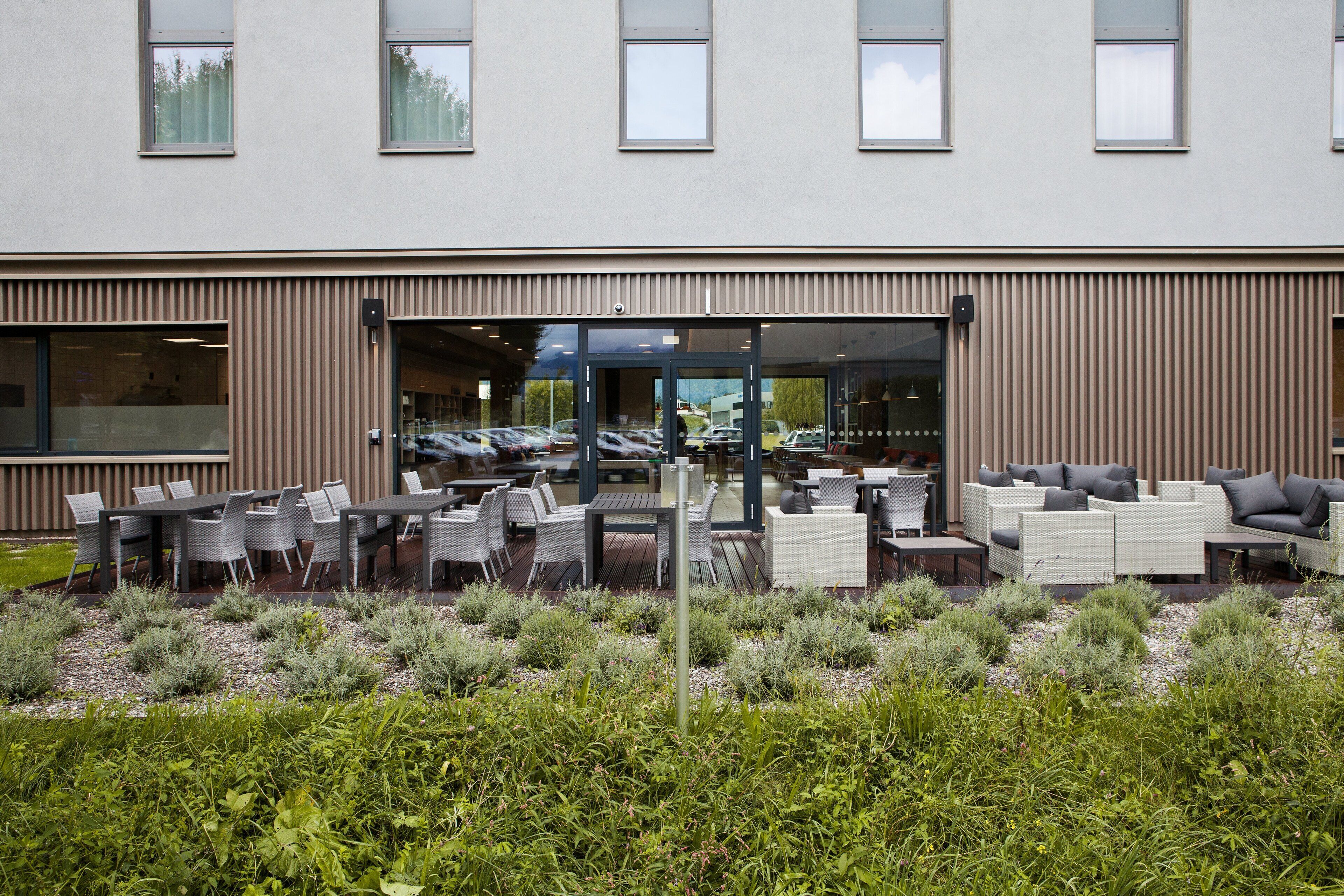 Un espace extérieur aménagé avec des tables et des chaises, l’entrée d’un bâtiment et un parking.