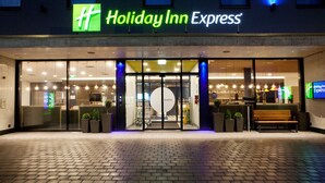The exterior of a Holiday Inn Express hotel with a glass entrance, illuminated signage, and a bench outside.