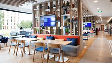 Een moderne hotel lobby met een zithoek, een groot scherm dat informatie toont, en uitzicht op de parkeerplaats.