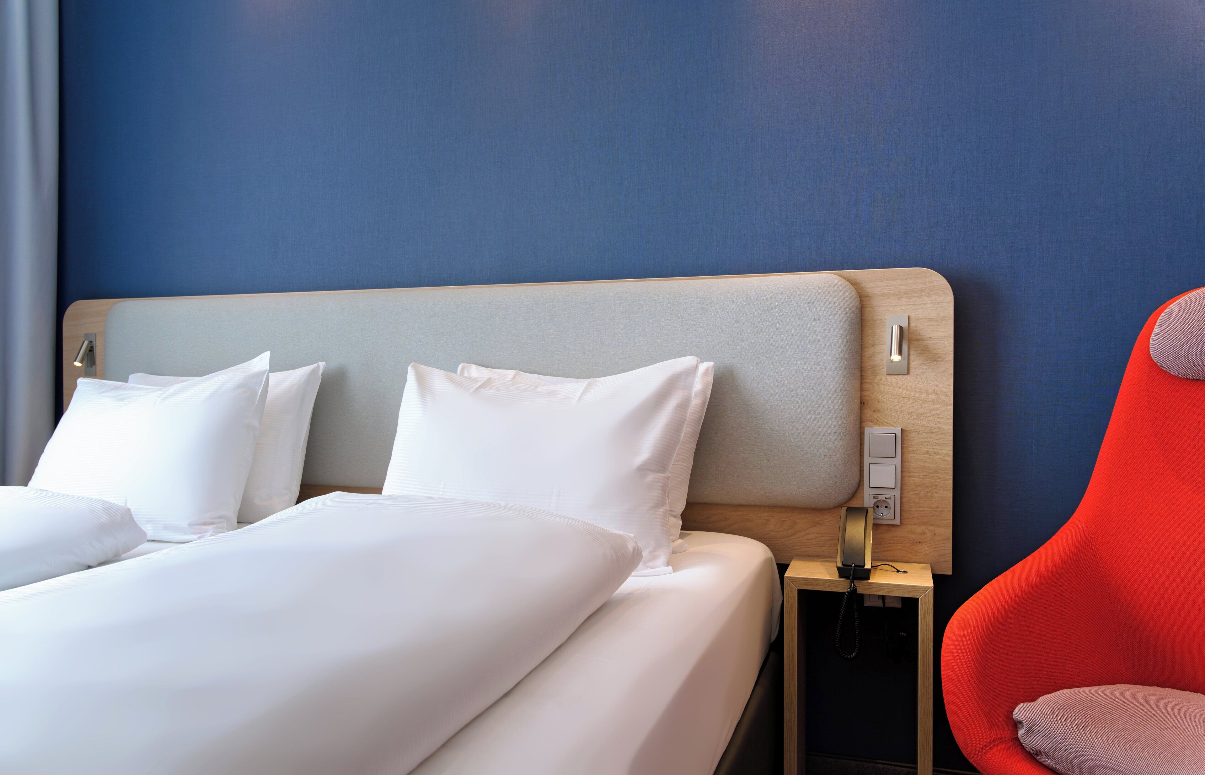 Quarto de hotel com cama, mesa de cabeceira, uma cadeira vermelha e interruptor na parede.