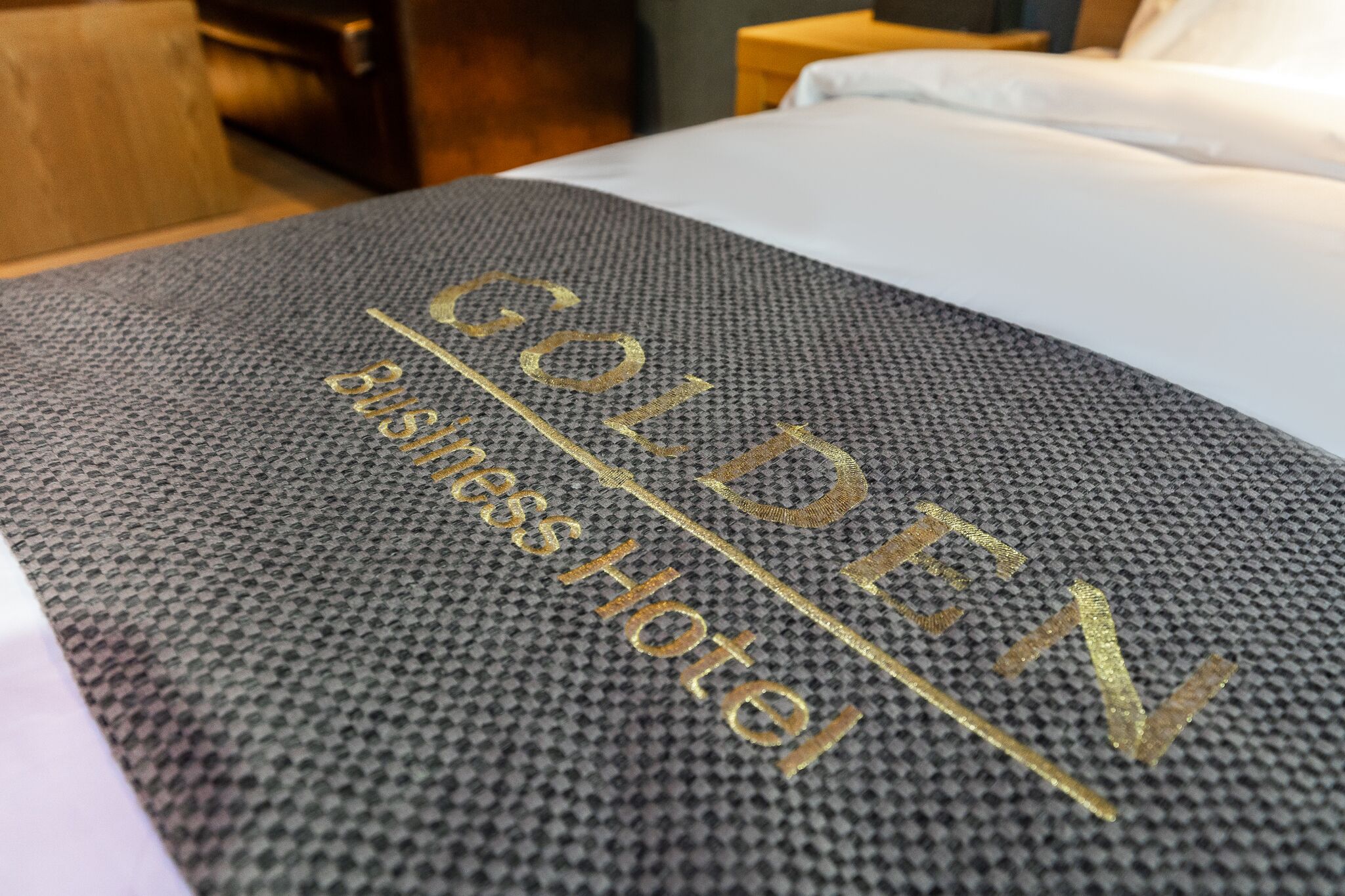 A bed with a textured gray bedspread and the text "GOLDEN BUSINESS HOTEL" embroidered in gold.