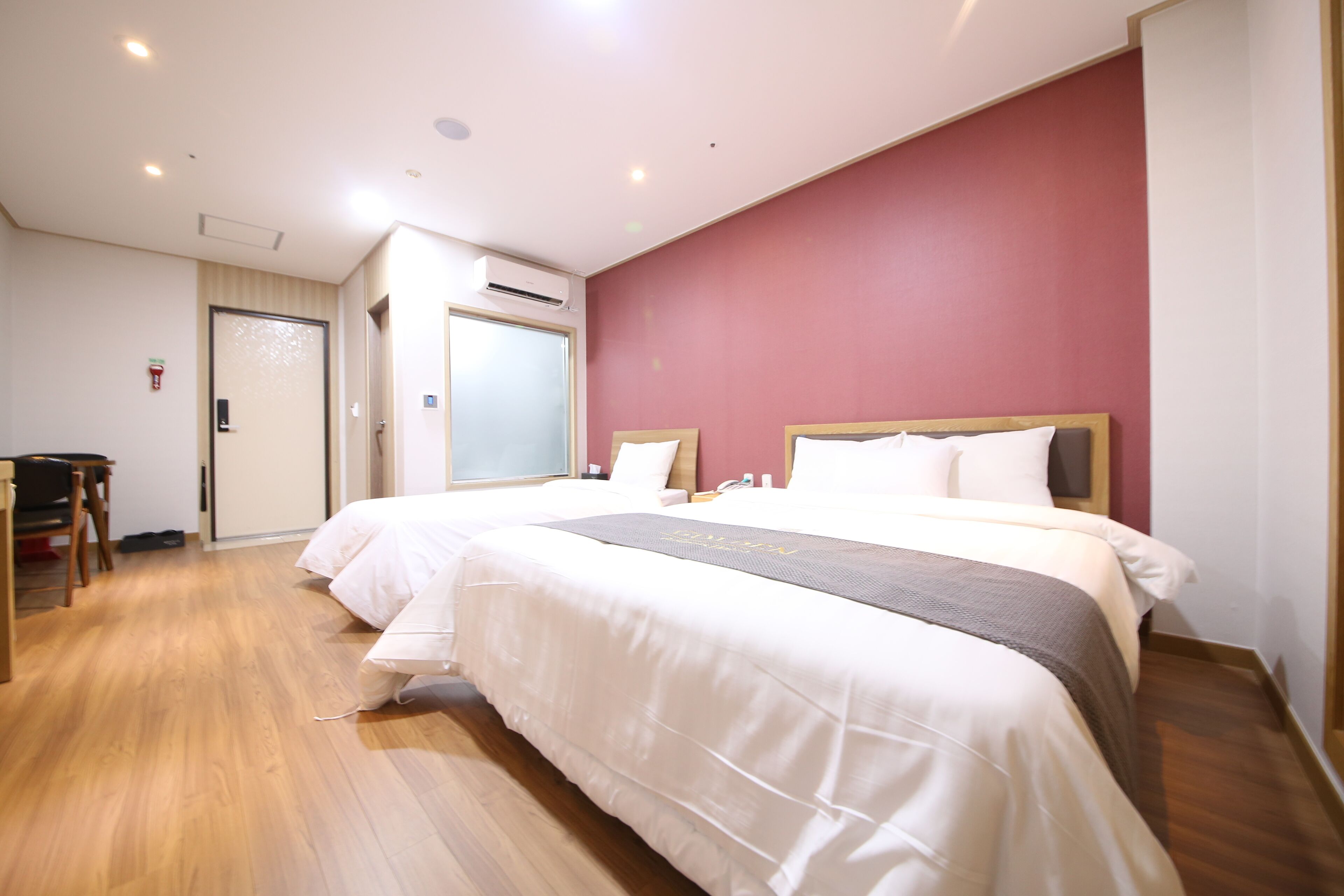 Quarto de hotel com duas camas, piso de madeira, parede com detalhes em vermelho e uma mesa de trabalho com cadeira.