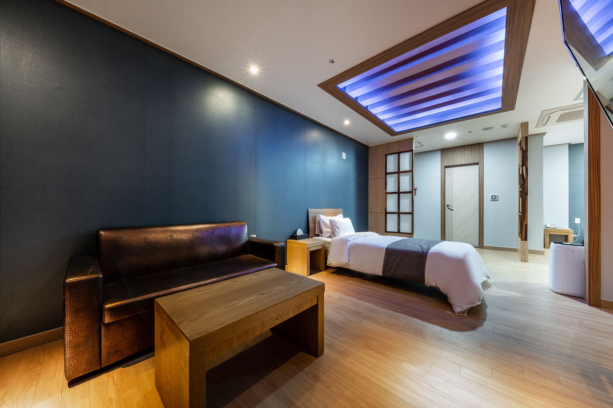 A modern hotel room with a dark blue accent wall, a wooden bench, a bed with white bedding, and a skylight with blue lighting.
