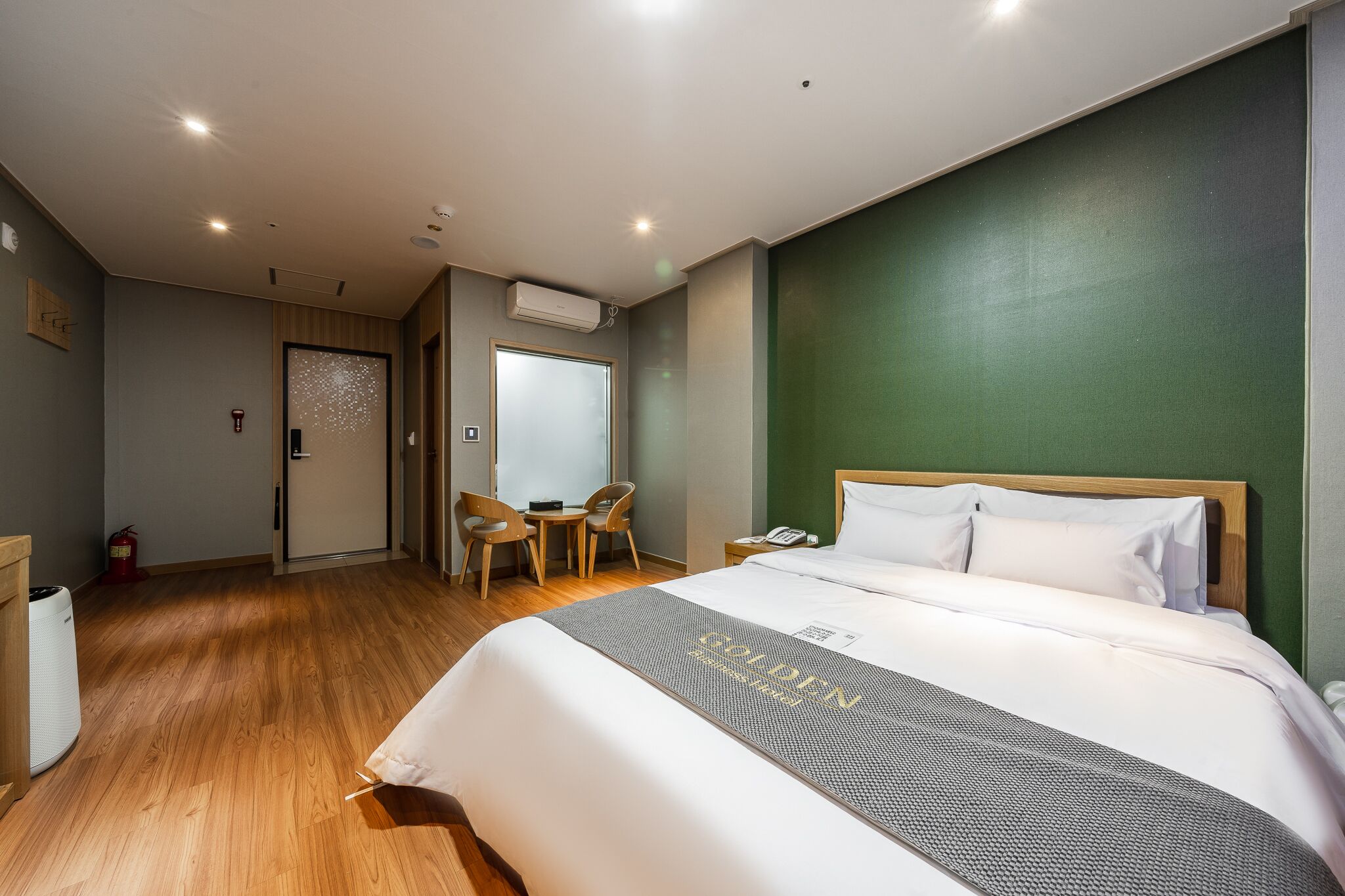 A hotel room with a large bed, a desk with a chair, a small table, and a green accent wall.