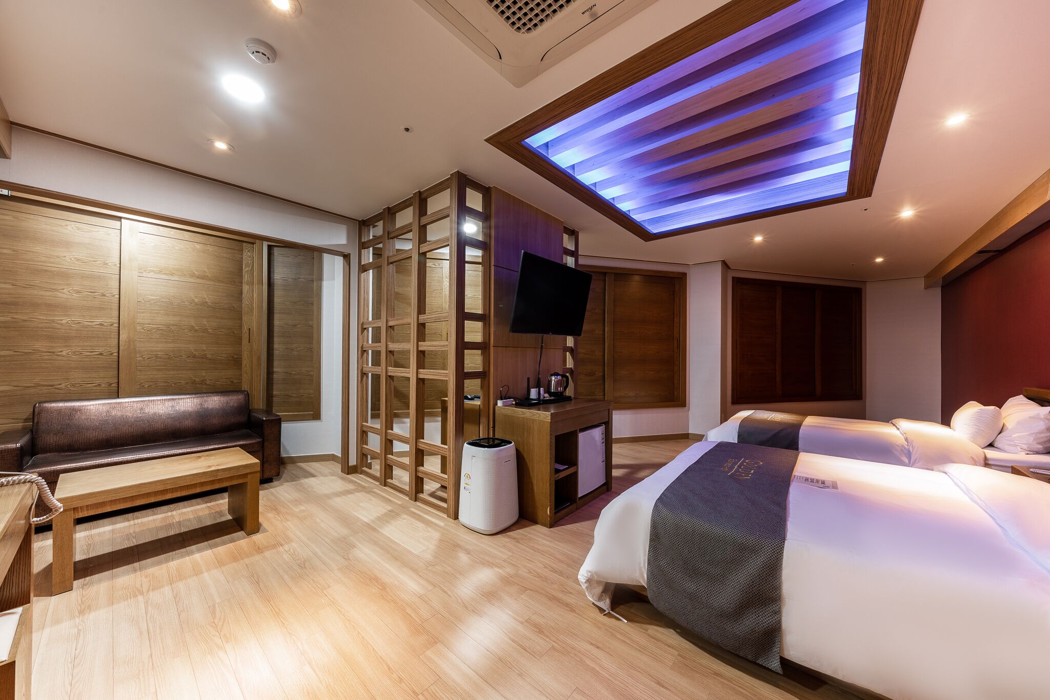 Quarto de hotel moderno com piso de madeira, sofá, cama com cobertor, escrivaninha com televisão de tela plana e teto com painel iluminado em azul.