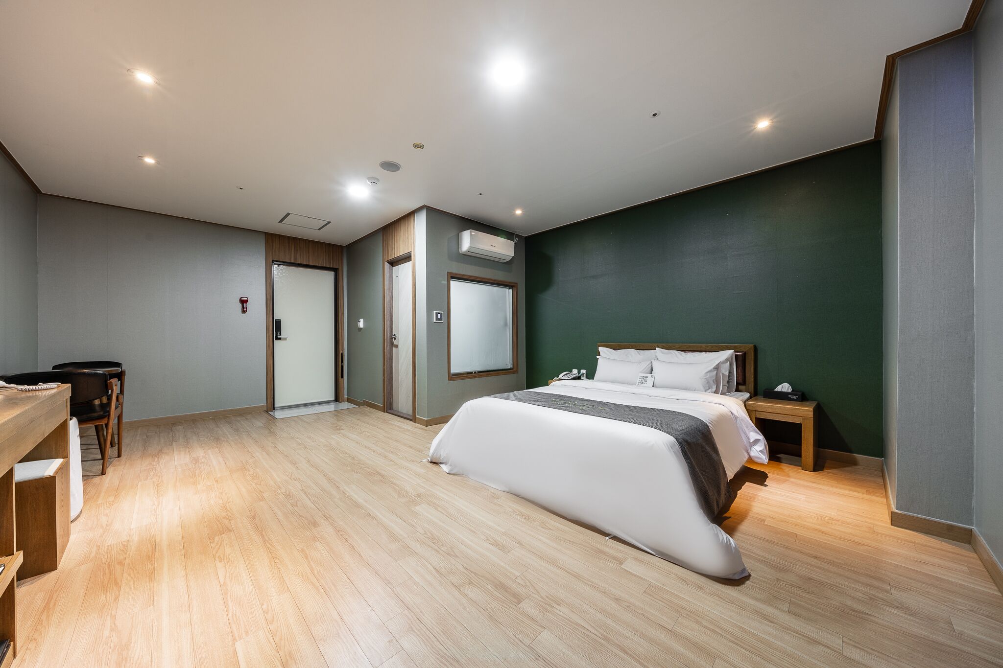 A modern hotel room with a large bed, a desk, and a chair. There is a door with a mirror and an air conditioning unit on the wall.