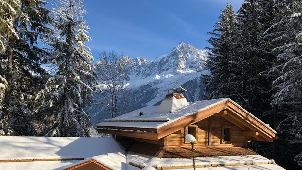 Una cabaña de madera con techo inclinado, rodeada de árboles nevados y montañas.