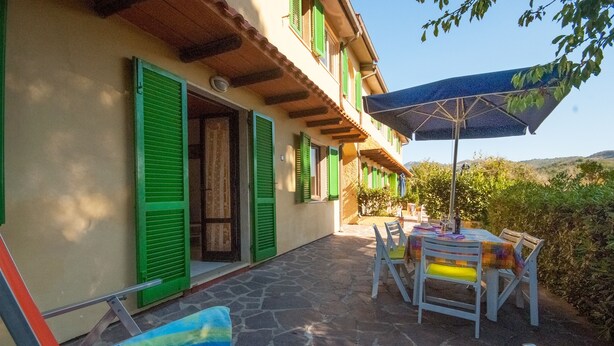 A patio with a table and chairs, a blue umbrella, and a building with green shutters. - Residence La Valdana - 3 room apartment with swimming pool, tennis and parking 1km from the beach (Capoliveri)