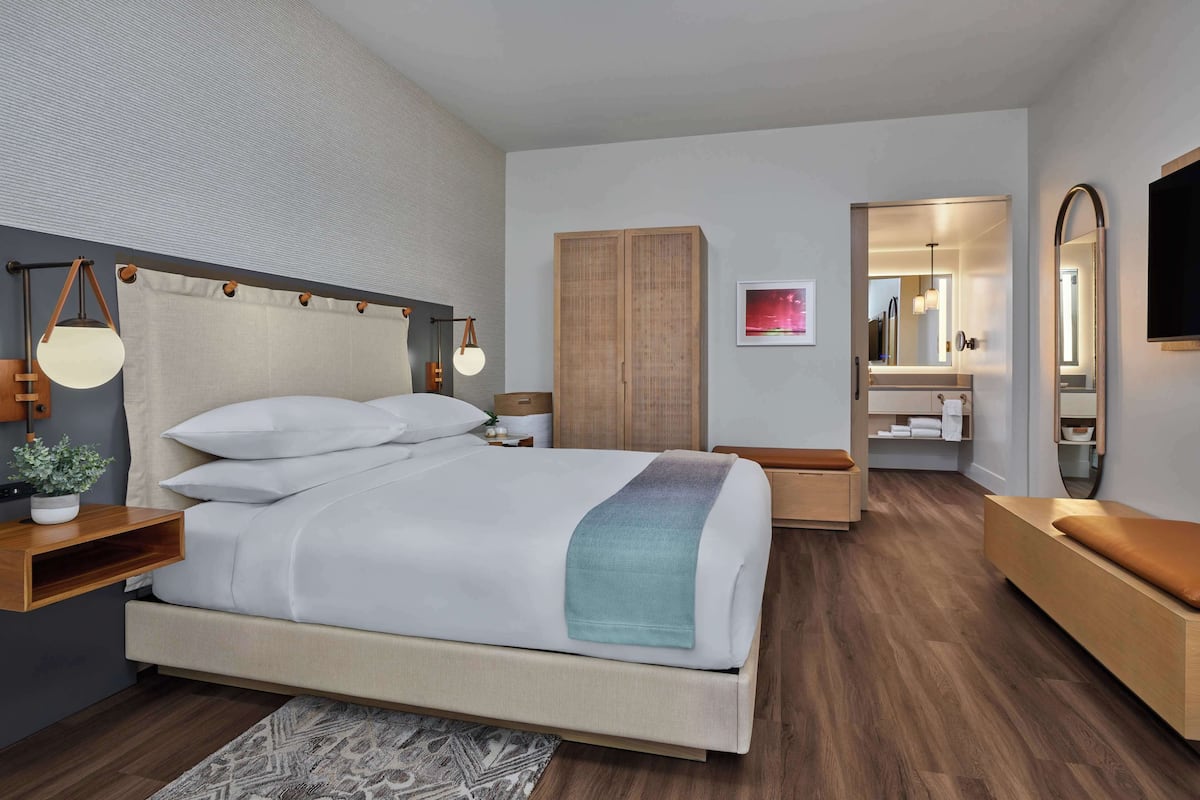 Una habitación de hotel moderna con una cama grande, mesitas de noche, un armario, un espejo y un televisor.