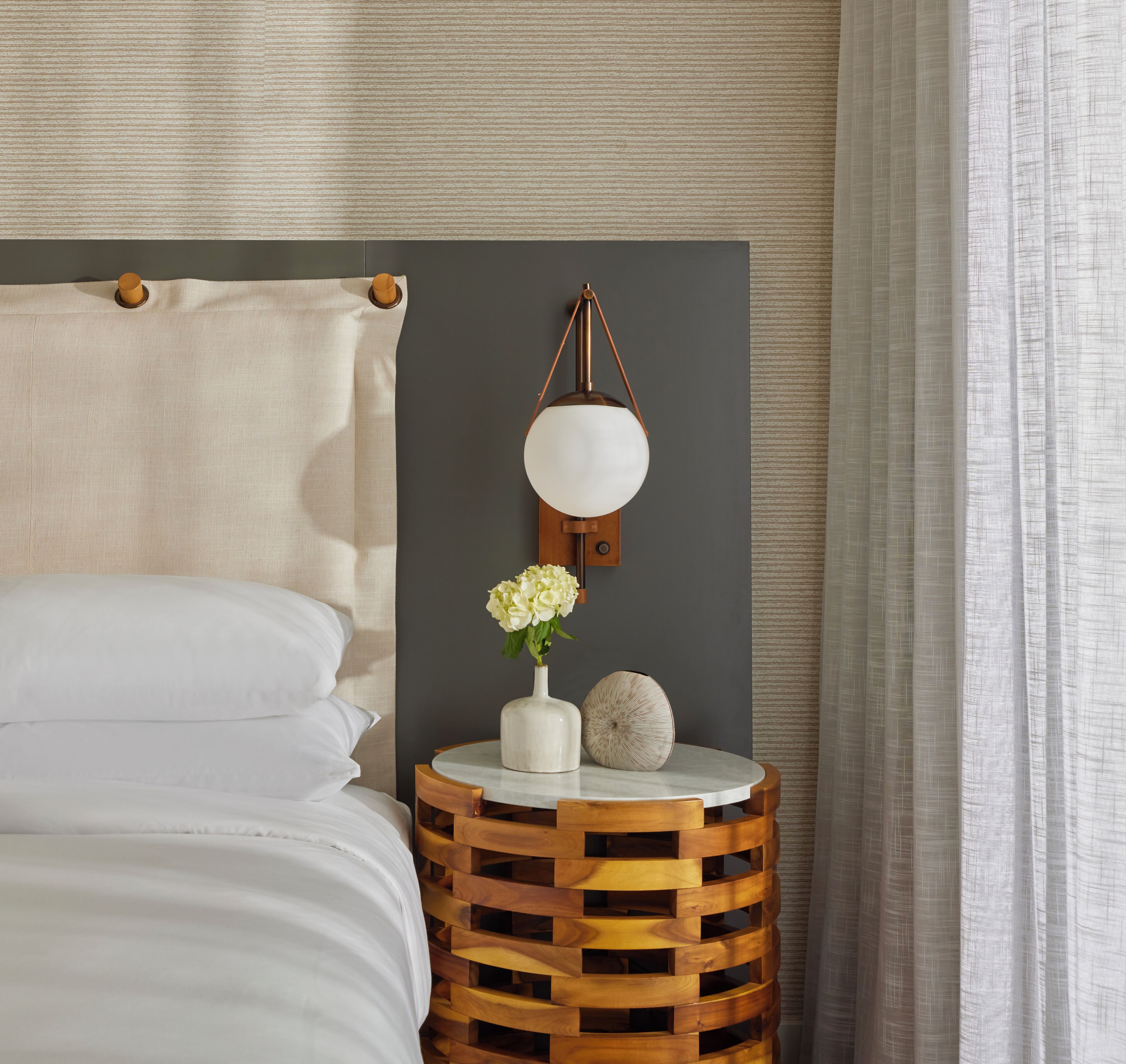 Uma cama bem arrumada com lençóis brancos, uma mesa de cabeceira redonda de madeira com um vaso branco e uma esfera decorativa, e um abajur fixado na parede.