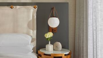 Uma cama bem arrumada com lençóis brancos, uma mesa de cabeceira redonda de madeira com um vaso branco e uma esfera decorativa, e um abajur fixado na parede.