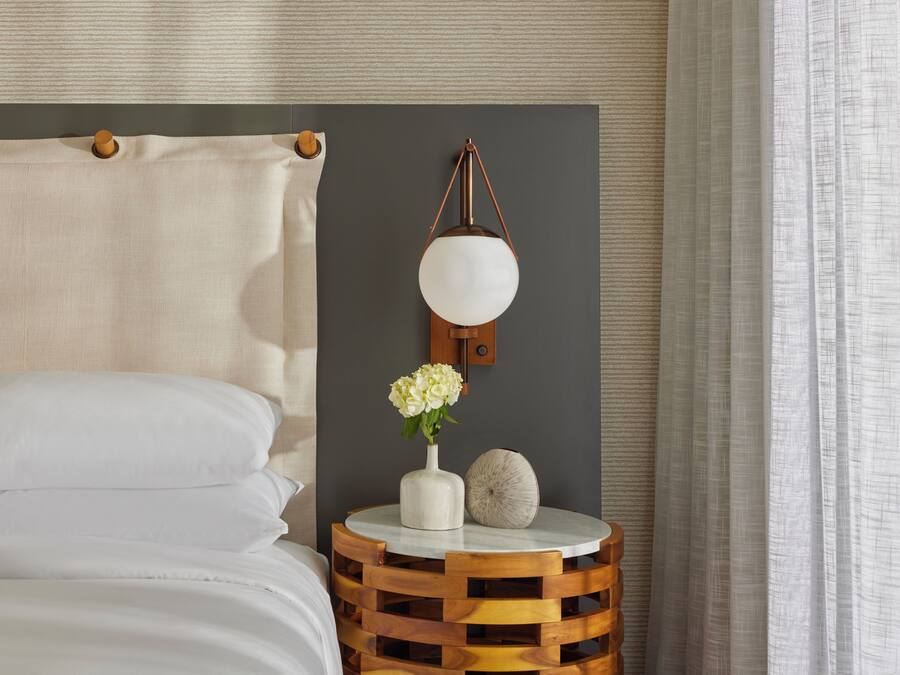 Uma cama bem arrumada com lençóis brancos, uma mesa de cabeceira redonda de madeira com um vaso branco e uma esfera decorativa, e um abajur fixado na parede.