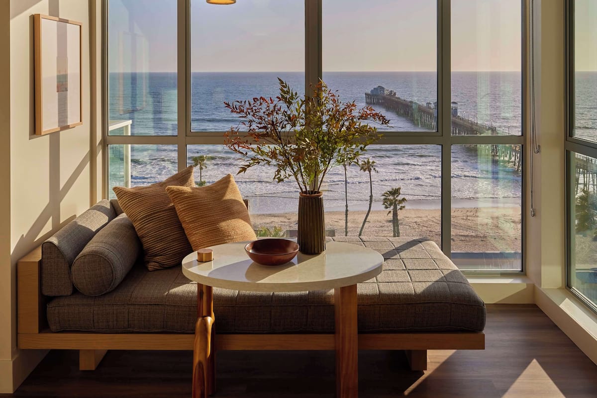Una sala de estar moderna con un amplio ventanal con vistas al océano, un sofá de madera, una mesa redonda y un colgador de luz.