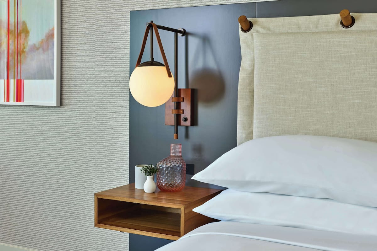 Una cama bien hecha con ropa blanca, una mesita de noche de madera con un jarrón de vidrio y una pequeña planta, y una lámpara de pared con pantalla redonda.