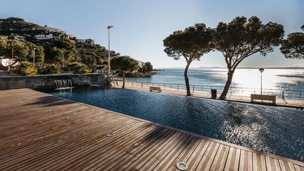 Une terrasse en bois avec une piscine, donnant sur une zone côtière bordée d’arbres et de bâtiments.