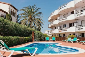 A swimming pool with lounge chairs, surrounded by greenery and a palm tree, with multi-story buildings in the background. - V005 - Cargols de Mar 5, Casa en residencia privada con piscina delante de la playa. (Empuriabrava)