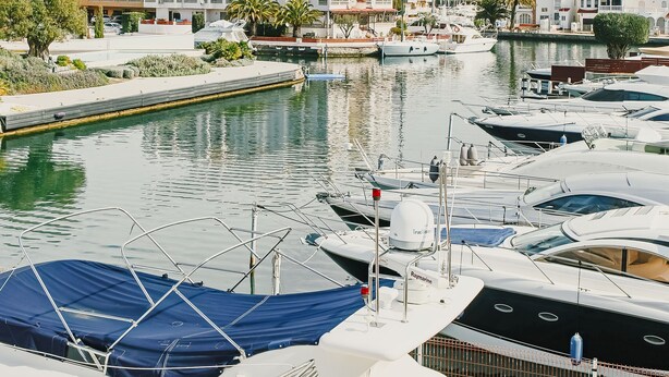 A marina with boats docked, surrounded by buildings with red-tiled roofs and a mountainous backdrop. - V005 - Cargols de Mar 5, Casa en residencia privada con piscina delante de la playa. (Empuriabrava)