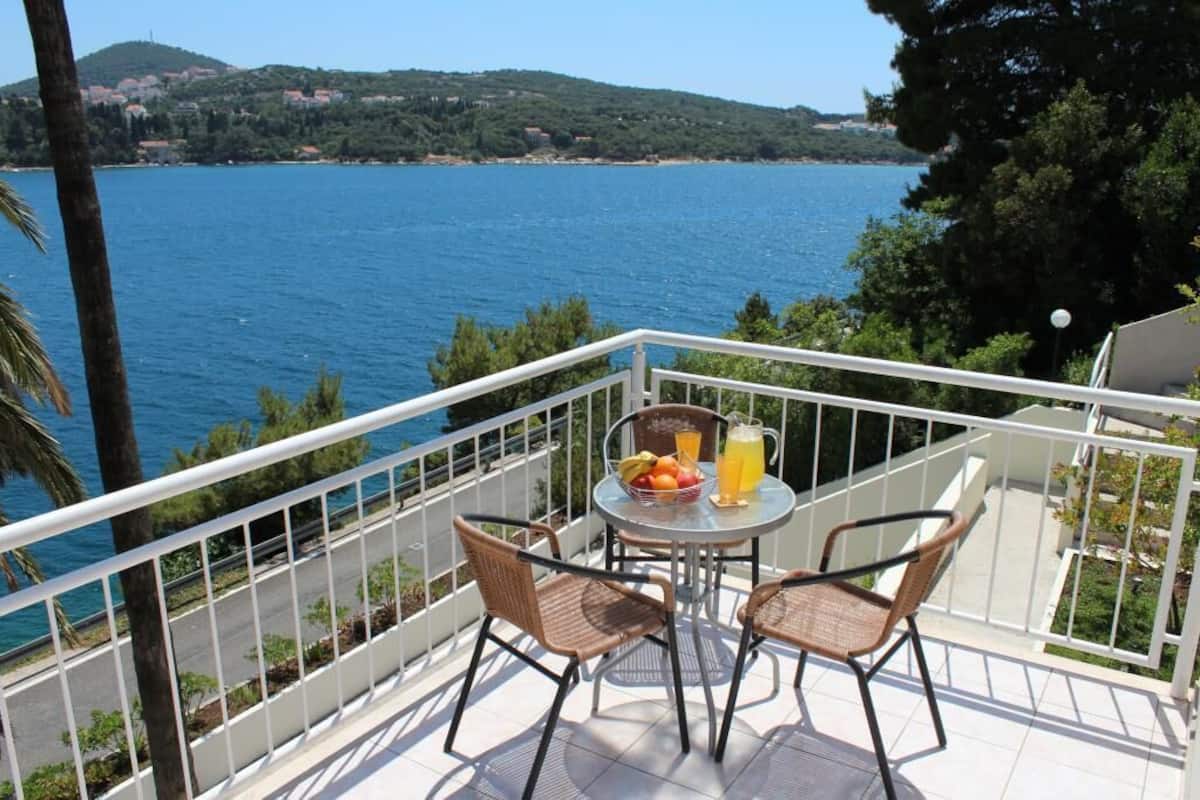 A balcony with wicker chairs and a table, overlooking a body of water and a distant coastline.
