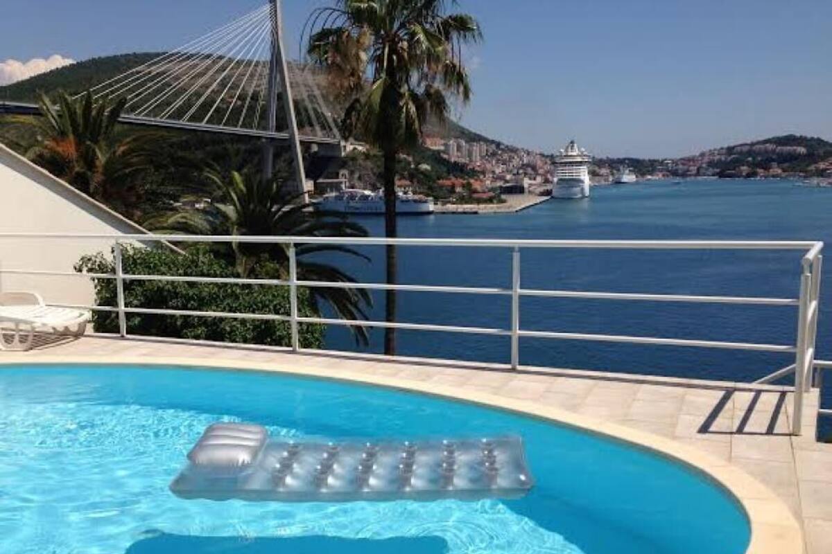 A swimming pool with an inflatable float, a palm tree, and a view of a coastal town with a bridge and a cruise ship.