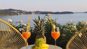Pöydällä on kaksi juomaa, ananas ja rypäleterttuja, ja taustalla avautuu kaunis merimaisema.