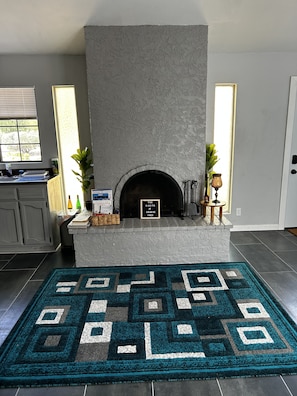 Una sala de estar con chimenea, un tapete estampado y una zona de cocina con armarios y ventana.
