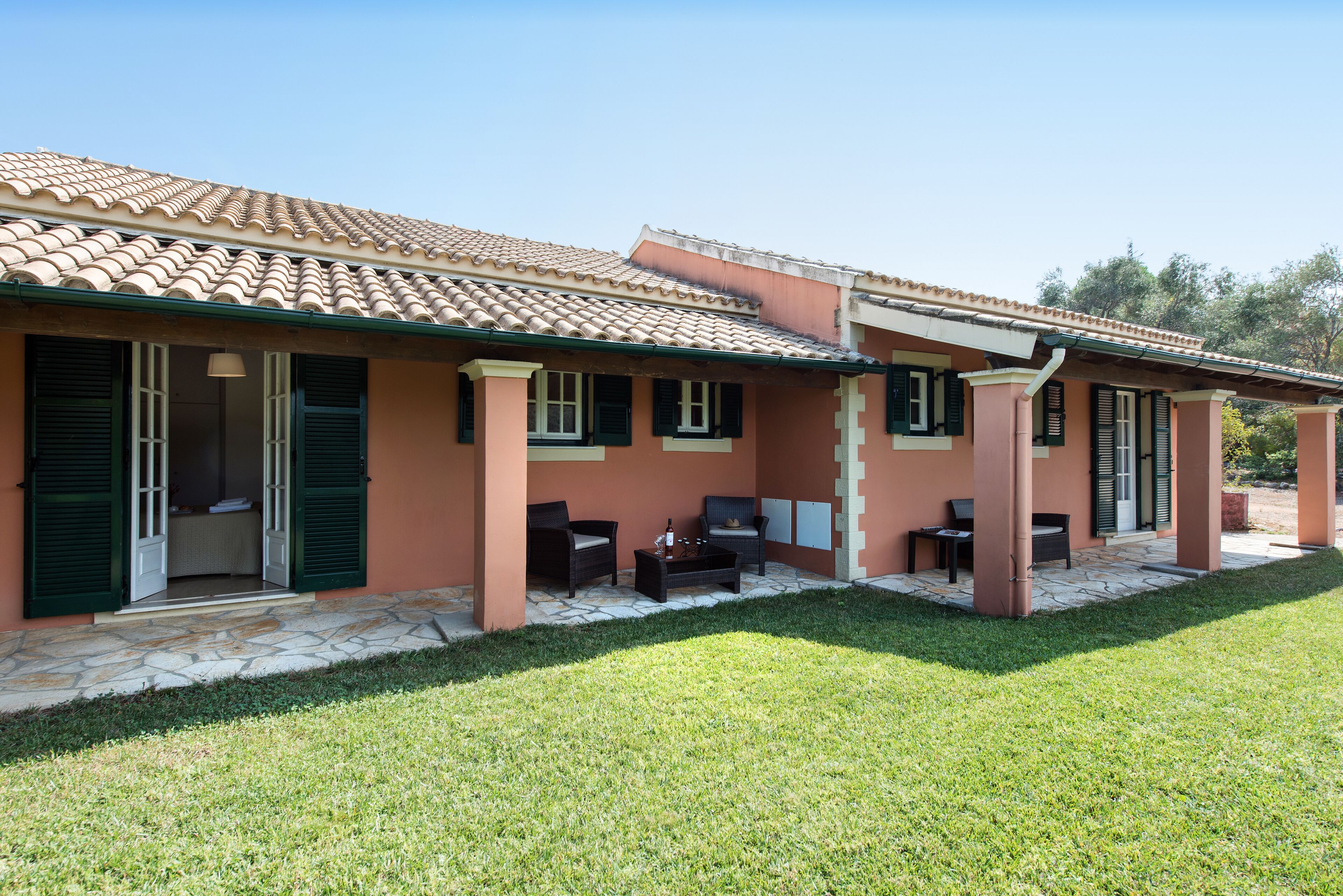 Una casa con techo de tejas, contraventanas verdes y un patio empedrado con mobiliario exterior.