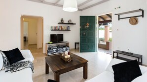 A living room with a sofa, a coffee table, a TV, and a bookshelf. There is a green door leading outside. - Villa Cesar (Corfu)